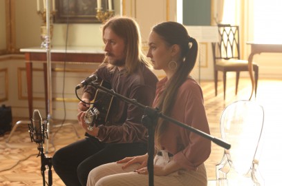 Mężczyzna z gitarą i kobieta siedzą na krzesłach przed mikrofonem.