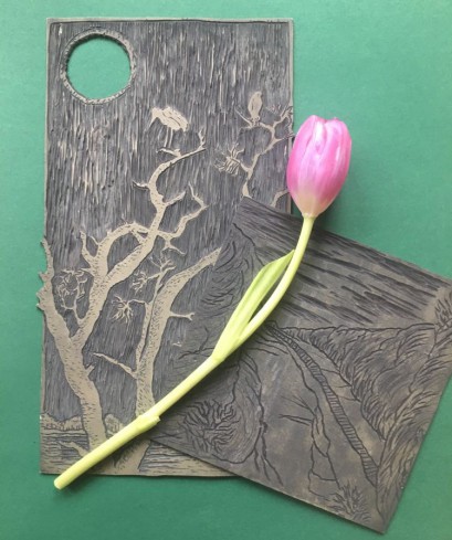 Różowy tulipan leżący na dwóch ciemnych drewnianych prostokątnych deseczkach. Deseczki są ozdobione płaskimi rzeźbieniami przedstawiającymi drzewa.