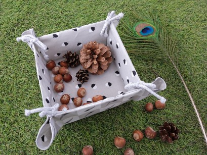 Kwadratowy biały koszyk z materiału stojący na trawie. W koszyku leżą szyszki, obok koszyka leży pawie pióro. 
