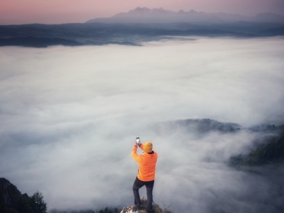 Mężczyzna robiący zdjęcie telefonem gór spowitych gęstą mgłą.