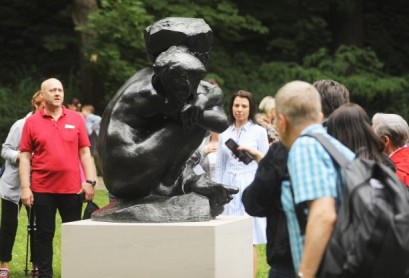Grupa ludzi stoi wokół czarnej rzeźby przedstawiającej skuloną postać.   
