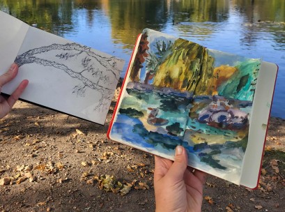 Kobiece dłonie trzymają dwa rysunki na tle wody w stawie: z lewej strony rysunek gałęzi wykonany ołówkiem, z prawej pejzaż namalowany farbami.