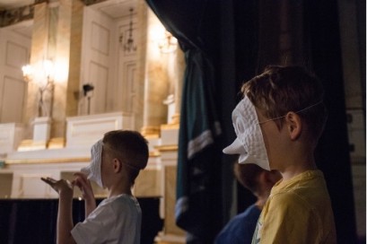 Dwóch chłopców w teatralnych maskach stoi za kurtyną.