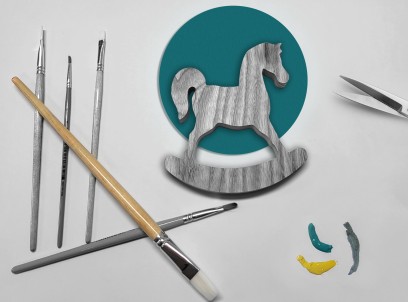 Pośrodku leży drewniany konik, po lewej jest kilka pędzelków, po prawej widać ostrze nożyczek. 