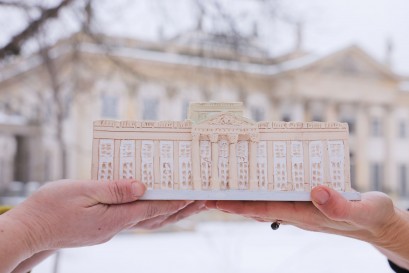 Kobiece dłonie trzymające ulepioną z gliny miniaturę Pałacu na Wyspie. Miniatura jest trzymana na tle budynku Pałacu na Wyspie. 