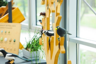 Cztery drewniane ptaki siedzące na drewnianej konstrukcji stojącej na stole. Obok, po lewej, stoi doniczka z kwiatem i makieta z ptasimi jajami.