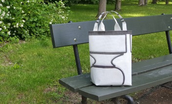 Fragment ławki w parku, na której stoi biała torba. 