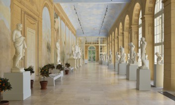 Galeria Rzeźby w Starej Oranżerii, pod oknem stoją ustawione na postumentach białe rzeźby, jako pierwsza widoczna jest rzeźba przedstawiająca nagich mężczyzn.