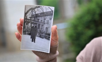 Dłoń kobiety trzymająca zdjęcie, na którym widać stojącego mężczyznę przed budynkiem. 