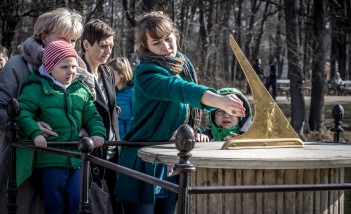 Trzy kobiety i dwoje dzieci stoją przed rzeźbą zegara słonecznego. Jedna z kobiet wskazuje ręką na zegar, który składa się z kamiennej tarczy i ustawionej na niej złotej wskazówki, uniesionej pod kątem do góry.