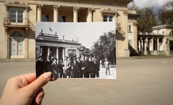 Kobieca dłoń trzymająca archiwalną fotografię na tle Pałacu na Wyspie. Na zdjęciu widoczna jest grupa ludzi pozująca na tle Pałacu na Wyspie. 