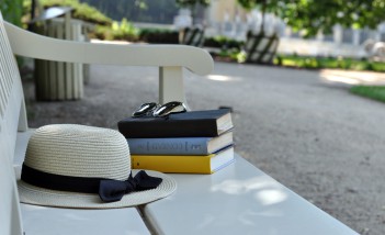 Parkowa ławka, na której leżą trzy książki, na książkach są okulary przeciwsłoneczne, a obok biały kapelusz przewiązany czarną wstążką.