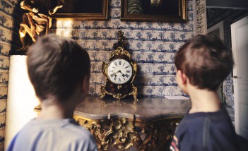 Dwóch chłopców stoi przed ścianą, na której wiszą obrazy. Na komodzie pod ścianą stoi zegar. 