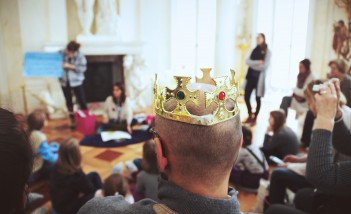 Na tle grupy dzieci i dorosłych zebranych w sali Pałacu na Wyspie widoczny jest stojący tyłem mężczyzna, który ma na głowie koronę. 