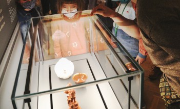 Kilkoro dzieci stoi przy szklanej, oświetlonej gablocie, w której są umieszczone trzy ceramiczne przedmioty. 