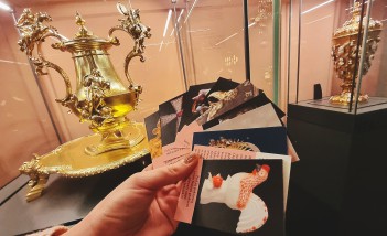 Kobieca ręka trzyma talię kolorowych kart na tle dwóch gablot wystawowych, w których znajdują się bogato zdobione złote wazony. 