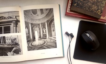 Otwarta książka ze zdjęciami obiektów Łazienek Królewskich, obok książki leżą słuchawki, podkładka z myszką oraz dwie inne, zamknięte książki. 