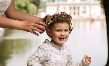 Dziewczynka z królewską koroną na głowie stoi na tle Pałacu na Wyspie. Kobiece dłonie dotykają korony na głowie dziecka.