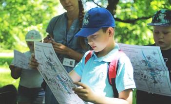 Kilkuletnie dzieci stoją w ogrodzie. W rękach trzymają mapy, na które patrzą.