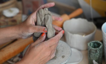 Kobiece dłonie ugniatające szary kawałek gliny nad spodkiem. Obok stoją ceramiczne kubki. 