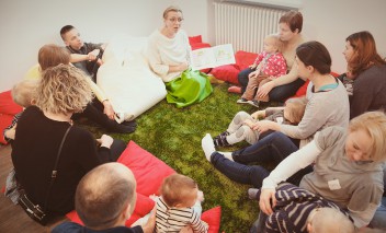 Grupa dorosłych z dziećmi siedzi na podłodze przed kobietą trzymającą otwartą książkę. 