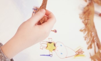 Ręka dziecka trzymająca kawałek materiału z frędzlami nad kartą z rysunkiem przedstawiającym postać. 
