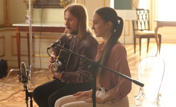 Mężczyzna z gitarą i kobieta siedzą na krzesłach przed mikrofonem.