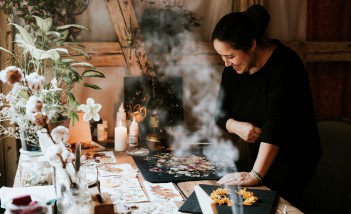 Kobieta ubrana na czarno pochyla się nad stołem, na którym leżą rozmaite gadżety, takie jak kwiaty, suche liście i kolorowe obrazki.