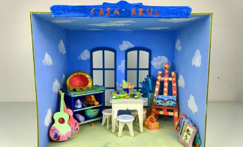 Wnętrze miniaturowego domku. W domku stoją oparte o ściany gitary, stół i trzy taborety, malarska paleta i obrazy oparte o prawą ścianę.
