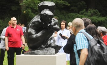 Grupa ludzi stoi wokół czarnej rzeźby przedstawiającej skuloną postać.   
