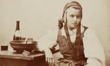 Dawna fotografia ukazująca dziewczynę w stroju ludowym siedzącą przy stole, na którym stoi miska, butelka i szklanka.