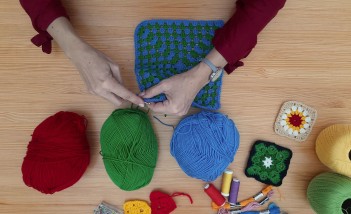 Widoczne są kobiece ręce, które robią na drutach. Obok leżą kolorowe włóczki i nitki.