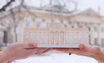 Kobiece dłonie trzymające ulepioną z gliny miniaturę Pałacu na Wyspie. Miniatura jest trzymana na tle budynku Pałacu na Wyspie. 