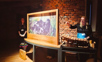 W kwadratowej, drewnianej ramie znajduje się plansza z ilustracją. Po obu stronach ramy stoją kobiety. Kobieta po prawej stronie uderza w drewniany instrument.