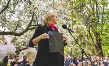Kobieta z czarną kartką trzymaną w rękach stoi przed mikrofonem. W tle widać tłum ludzi stojący pod drzewami.