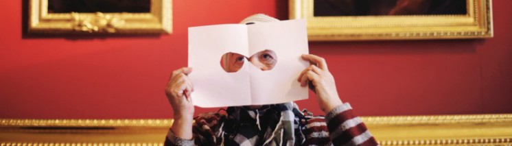 Kobieta trzyma kartkę papieru z wyciętymi otworami, którą przykłada do twarzy, w tle, za jej głową, wiszą obrazy.