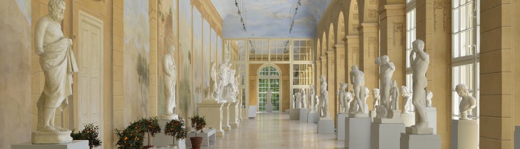 Galeria Rzeźby w Starej Oranżerii, pod oknem stoją ustawione na postumentach białe rzeźby, jako pierwsza widoczna jest rzeźba przedstawiająca nagich mężczyzn.