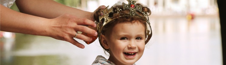 Dziewczynka z królewską koroną na głowie stoi na tle Pałacu na Wyspie. Kobiece dłonie dotykają korony na głowie dziecka.