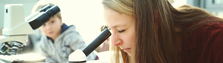 Kobieta patrząca przez mikroskop, za kobietą, na stole, stoi kolejny mikroskop, przy którym siedzi małe dziecko.